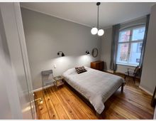 Schlafzimmer mit hellem Holzboden und grauen Wänden. Ein Doppelbett mit grauer Tagesdecke steht unter einer Wand mit zwei schwarzen Leselampen, daneben ein kleiner Stuhl und ein Beistelltisch. Ein großes Fenster mit grauen Vorhängen bietet Blick nach draußen, ergänzt durch eine hölzerne Kommode und einen Stuhl.