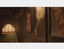 Ein stimmungsvoll beleuchteter Flur mit dunkelgrünen Wänden, an denen Porträts in Holzrahmen hängen. Links steht eine klassische Stehlampe mit einem weißen, geschwungenen Schirm und Fransen, metallene Rohre verlaufen entlang der Decke.