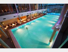 Innenansicht eines modernen Hallenbades mit einem großen rechteckigen Schwimmbecken. Rund um das Becken stehen mehrere Liegen, und durch eine Glaswand ist eine Außenlandschaft sichtbar.