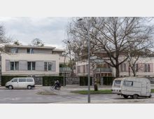 Moderne Wohnhäuser mit Flachdächern, großen Fenstern und horizontalen Fassadenelementen, davor eine Straße mit parkenden Fahrzeugen, darunter ein Wohnwagen und ein Kleinbus, sowie ein Radfahrer. Im Vordergrund stehen Straßenschilder und einige kahle Bäume.