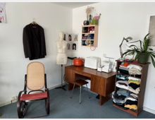 Small sewing and craft studio corner with a wooden desk holding a serger and covered machine, a dress form beside it, and wall-mounted cubbies filled with yarn. A rocking chair, a hanging jacket, a shelving unit stacked with folded fabric, and a desk lamp with potted plants sit against white walls on a carpeted floor.