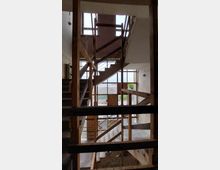 Treppenhaus in einem Rohbau mit freiliegendem Holz- und Metallgeländer, roh verputzten Wänden und großen Fenstern, die Tageslicht hineinlassen. Im Hintergrund sind Außenbereiche und Vegetation sichtbar.