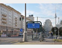Straßenansicht des Strausberger Platzes in Berlin, mit einem U-Bahn-Schild im Vordergrund. Im Hintergrund sind klassische DDR-Architekturbauten und der Fernsehturm zu sehen, daneben belebt durch Autos, Fahrradfahrer und Straßenverkehrsschilder.