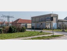 Straßenszene in einem Gewerbegebiet mit einem flachen Gebäude aus grauem Putz, das Graffiti aufweist, und einem benachbarten Gebäude mit rotem Ziegeldach. Im Vordergrund befindet sich eine Grünfläche mit Gehwegen, mehreren Straßenschildern und einem Strommast im Hintergrund.