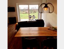 Wohnzimmer mit Esstisch aus Holz, im Hintergrund eine große Glastür, die den Blick auf einen Garten mit Rasen und Bäumen freigibt. Eine Hängeleuchte aus Metall und eine schwarze Couch sind Teil der Einrichtung.