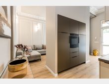 Offener Wohn- und Küchenbereich mit hellem Holzboden, weiß vertäfelten Wänden und Stuckdecke. Links steht ein graues Ecksofa im Wohnbereich, rechts eine moderne dunkle Einbauküchenwand mit integriertem Backofen neben einer Glastür zum Außenbereich.