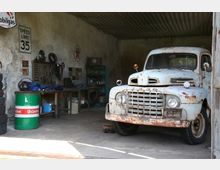 Eine alte Autowerkstatt mit einer rostigen, weißen Oldtimer-Lieferwagenfront, die aus einem offenen Tor herausragt. Im Hintergrund sind Werkbänke mit Werkzeugen, Regale, Reifen und dekorative Schilder an der Betonwand zu sehen.