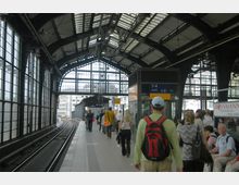 Bahnhofshalle mit hohen Stahlträgern und großen rechteckigen Fensterfronten, einem Gleis auf der linken Seite und einer überdachten Bahnsteigplattform, auf der mehrere Personen gehen oder warten. Ein Schild weist Richtung Ausgang sowie auf barrierefreie Einrichtungen hin.
