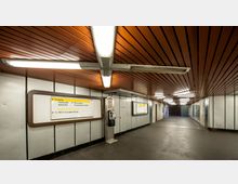 Unterirdischer U-Bahn-Eingangsbereich mit einer Decke aus hölzernen Paneelen und heller Beleuchtung. An den Wänden befinden sich Richtungsschilder und ein öffentlicher Telefonapparat, und der Boden ist aus dunklem Beton.