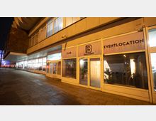 Außenansicht eines modernen Veranstaltungsortes bei Nacht, mit großen Fenstern und einer Beschilderung mit der Aufschrift „Balcony Club Eventlocation“. Das Gebäude hat eine angestrahlte Fassade und daneben eine überdachte Gehwegfläche.