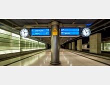 Eine moderne, beleuchtete Bahnhofshalle mit glatten Böden, einer zentralen Säule, digitalen Zuganzeigetafeln und analogen Uhren. Links befindet sich eine Wand aus satiniertem Glas, und rechts sind Betonpfeiler sichtbar.