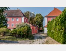 Ein schmaler, gepflasterter Weg führt durch eine ländliche Szene mit roten Wohnhäusern im klassischen Stil, umgeben von grünen Hecken, Bäumen und dichter Vegetation. Die Dächer der Häuser sind mit roten Ziegeln gedeckt, während ein einfacher Holzzaun und eine Straßenlaterne das Bild ergänzen.