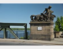 Steinfigur eines Mannes mit Pferd auf einem Sockel an einer Brücke, daneben ein grünes Brückenelement aus Stahl. Im Hintergrund ist ein Fluss mit Bäumen und einem klaren Himmel zu sehen.