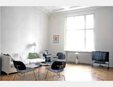 Helles Wohnzimmer mit weiß gestrichenen Wänden, einem großen Fenster mit Sprossen, einem Sofa mit grünen Kissen, schwarzen Stühlen und einem kleinen Couchtisch. Der Raum hat Parkettboden, einen Heizkörper unter dem Fenster und einen Fernseher auf einem Rollständer.