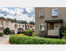 Reihenhaussiedlung mit zwei- bis dreistöckigen Häusern, Putzfassaden in beige und weiß, roten und schwarzen Ziegeldächern sowie kleinen Vorgärten mit Hecken und Pflanzen. Im Vordergrund ein Hauseingang mit Vordach und Treppe, im Hintergrund vereinzelte Bäume und Wolken am Himmel.