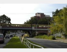 Straßenszene mit einer gelben S-Bahn, die über eine Brücke fährt, darunter eine mehrspurige Straße mit Autos. Im Hintergrund sind Bäume und ein modernes Backsteingebäude zu sehen, der Gehweg ist von einer Wiese mit Geländer gesäumt.