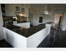 Moderne Küche mit weißen Schränken, glänzenden schwarzen Granit-Arbeitsplatten und integrierten Geräten. Die Küche verfügt über eine zentrale Kochinsel mit einer Dunstabzugshaube aus Edelstahl und zwei Barhockern vor einer erhöhten Theke.