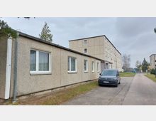 Ein einfacher, mehrstöckiger Wohnblock aus hellbraunem Beton mit rechteckigen Fenstern, flankiert von einem niedrigeren Anbau. Im Vordergrund steht ein grauer Lieferwagen auf einer schmalen gepflasterten Straße. Im Hintergrund sind Bäume und ein weiterer Betonbau zu sehen.