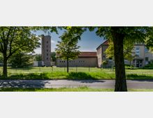 Straßenszene mit Blick auf ein eingezäuntes Grundstück, das aus mehreren beigefarbenen Gebäuden mit roten Dächern und einem hohen Turm besteht, umgeben von Bäumen und Grünflächen. Im Vordergrund verläuft eine Straße, die teilweise von Schatten der Bäume bedeckt ist.