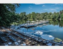 Lakeside beach area with rows of lounge chairs and white umbrellas on a sandy shore, next to floating dock platforms with cabanas extending onto a calm lake. Dense green trees surround the water under a clear blue sky, with reflections visible on the lake surface.