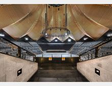 Moderner Konzert- oder Veranstaltungssaal mit einer zentralen Bühne, umgeben von ansteigenden Sitzreihen. Die Decke ist mit wellenförmigen, holzfarbenen Paneelen gestaltet, die den Raum akustisch und visuell prägen.