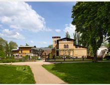 Historisches Gebäudeensemble in einer parkähnlichen Umgebung, bestehend aus einem ockergelben Haus mit Satteldach und Rundbögen sowie angeschlossenen Pergolen. Ein Kiesweg führt durch die gepflegte Rasenfläche, die von Bäumen und Ziersträuchern gesäumt ist.