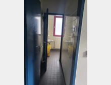 Schmaler Flur mit gefliesten Wänden und dunklem Boden, der zu einem kleinen Badezimmer mit einem Waschbecken, einer gelben Mülltonne und einem Fenster mit rotem Rahmen führt.