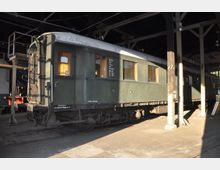 Ein historischer grüner Eisenbahnwagen der zweiten Klasse in einem überdachten Lokschuppen. Der Wagen hat große Fenster mit Vorhängen, sichtbare technische Details wie Leitungen und eine Beschriftung mit technischen Daten an der Seite.