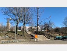 Städtischer Park mit kahlen Bäumen im Winter, einer breiten Steintreppe mit Geländern und einem orangefarbenen Mülleimer in der Mitte. Im Hintergrund ist ein modernes, klotzartiges Gebäude sowie einige Hochhäuser sichtbar, umgeben von einer Rasenfläche und Gehwegen.