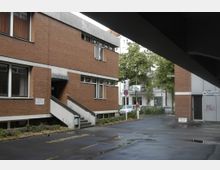 Zweigeschossiges Backsteingebäude mit der Aufschrift „Haus der Kirche“ und einem Treppeneingang an der Seite. Im Hintergrund befinden sich Bäume, ein weißes Wohngebäude sowie ein Parkplatz mit Autos.