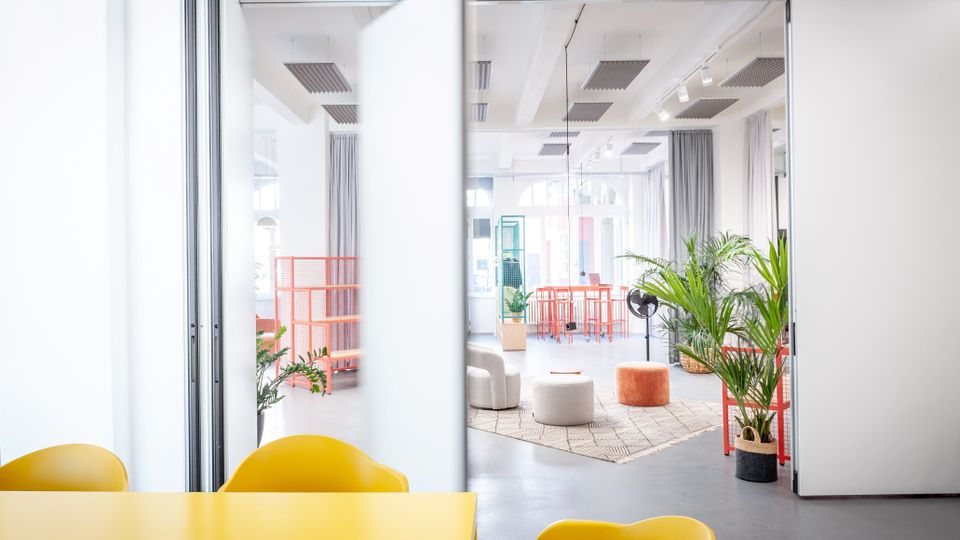Heller, moderner Büroraum mit großen Glas-Schiebetüren, hohen Decken mit Akustikpaneelen und viel Tageslicht. Im Hintergrund stehen orange Regale und Tische, mehrere Pflanzen sowie runde Sitzhocker auf einem Teppich; vorne ist ein gelber Tisch mit Stühlen zu sehen.