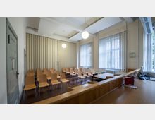 Ein kleiner Konferenzraum mit gestaffelt aufgestellten Holzstühlen, einem schlichten Schreibtisch mit Büromaterialien und Fenstern mit vertikalen Jalousien. Die Wände sind beige, und die Decke ist mit weißen Akustikplatten ausgestattet, während eine faltbare Trennwand im Hintergrund zu sehen ist.