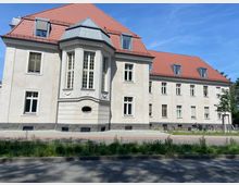 Zweistöckiges Gebäude mit heller Fassade, roten Ziegeldächern und einem mittig vorspringenden Erker mit großen Fenstern. Vor dem Gebäude befinden sich eine Straße, ein schmaler Streifen mit Grünpflanzen und ein Gehweg.