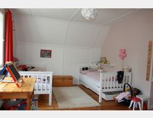 Kinderzimmer mit zwei weißen Einzelbetten, jeweils mit rosa Bettwäsche. Ein Holztisch mit einer kleinen Kreidetafel steht links, während eine Holzkiste und ein Teppich in der Mitte des Raumes liegen. Spielzeug und Bücher befinden sich an verschiedenen Stellen, darunter ein Regal an der Wand und eine Spielzeugbox unter einem Bett. Wände sind in Weiß und Rosa gehalten, mit einem Dachschrägen-Design.