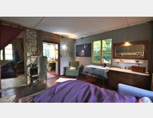 Gemütlicher Wohnbereich mit Holzboden und rustikalem Steinofen in der Mitte. Der Raum verfügt über Holzmöbel, ein großes Fenster mit Blick ins Grüne, eine Sitzecke mit Sessel sowie einen angrenzenden Arbeitsbereich.