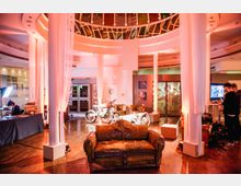 Innenraum eines modernen Gebäudes mit einer kreisförmigen Glaskuppel, weißen Säulen und rot-orangefarbener Beleuchtung. Im Vordergrund steht ein abgenutztes Ledersofa, dahinter sind zwei Motorräder ausgestellt, und an den Seiten befinden sich Arbeitsbereiche und Dekorationen.