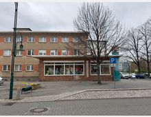 Zweistöckiges Gebäude mit beigefarbener und roter Klinkerfassade, im Erdgeschoss ein Geschäft mit Schaufenstern. Im Vordergrund ein Gehweg mit Pflastersteinen, ein Baum ohne Laub, ein Parkverbotsschild und eine kleine bepflanzte Fläche.