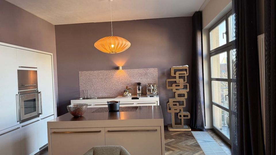 Modern kitchen interior with white built-in cabinets, a central island with a dark countertop, and a hanging pendant light against gray walls. Large floor-to-ceiling windows with dark curtains line one side, and a tall sculptural shelving unit stands near the window on a herringbone wood floor.