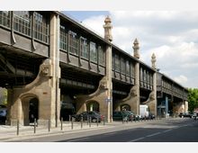 Erhöht verlaufende historische Eisenbahnbrücke mit sichtbaren steinernen Pfeilern und Metallkonstruktion, darunter eine Straße mit parkenden Autos und Fußgängerbereich. Im Hintergrund sind Elemente des Stadtbilds und ein bewölkter Himmel erkennbar.