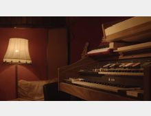 Ein gemütlicher Raum mit gedämpfter Beleuchtung, ausgestattet mit einer Stehlampe, einem Sofa und einem alten elektronischen Keyboard mit mehreren Schaltern und Tasten. Im Hintergrund sind eine Gitarre und rotbraune Wände sichtbar.