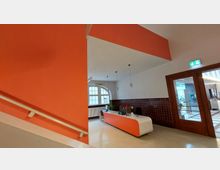 Empfangsbereich eines Gebäudes mit orangefarbenen Wänden und hellweißem Boden. Ein geschwungener Empfangstresen in Weiß und Orange steht vor einer halbhohen braunen Kachelwand, neben einem großen Fenster mit bogenförmigem Rahmen; eine Tür mit Glaseinsatz führt zu einem angrenzenden Bereich.