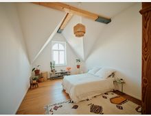 Schlafzimmer mit schräger Decke und einem großen Fenster mit Rundbogen. Der Raum hat Holzboden, ein schlichtes Bett mit weißer Bettwäsche, eine helle Hängelampe aus Rattan und mehrere Pflanzen sowie ein niedriger Regal mit Büchern und Dekoration.
