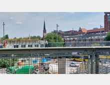 Eine Stadtansicht mit einer erhöhten Fußgängerbrücke im Vordergrund, auf der mehrere Personen laufen. Im Hintergrund sind eine Kirche mit einem spitzen Kirchturm, Backsteingebäude und ein modernes Gebäude mit großen Fenstern sichtbar. Darunter befinden sich Baustellenfahrzeuge und ein Busbahnhof.