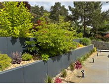 Eine moderne Gartengestaltung mit erhöhten Beeten aus grauen Betonwänden, gefüllt mit verschiedenen Pflanzen und Sträuchern. Der Boden ist mit Pflastersteinen belegt, und im Hintergrund sind Bäume und eine Mauer aus Naturstein sichtbar.