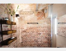 Badezimmer mit sichtbaren Ziegelwänden, einem kleinen Holzregal an der linken Seite mit dekorativen Objekten und einem schmalen Handtuchhalter aus Metall. Ein Spiegel mit beleuchtetem Rand und eine Deckenbelüftung sind ebenfalls sichtbar.