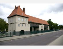 Gebäude im traditionellen Stil mit hellbeiger Fassade, roten Ziegeldächern und einem Turm mit mehreren kleinen Fenstern; im Vordergrund befindet sich eine Straße mit einem grünen Metallgeländer, im Hintergrund stehen Bäume und weitere Gebäude.