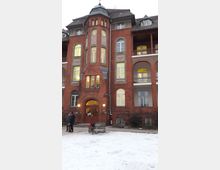 Backstein-Gebäude mit drei Etagen, zentralem Rundturm und Fenstern mit Bögen im historischen Stil. Vor dem Gebäude liegt ein schneebedeckter Platz, auf dem eine Bank und ein Fahrrad stehen.