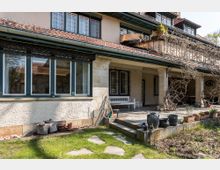 Eine gepflegte Terrasse eines Hauses mit heller Putzfassade und dunklem Satteldach. Die Terrasse ist mit weißen Säulen, einer weißen Bank und Pflanzgefäßen gestaltet; im Vordergrund befinden sich ein kleiner Garten mit Rasen und ein gepflasterter Weg.