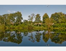 Ruhiger See mit gespiegelten Bäumen am Ufer, dahinter dichtes Grün und vereinzelte weiße Häuser mit roten Dächern. Am Himmel fliegt ein Flugzeug über die Szenerie.