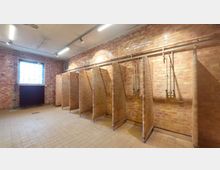 Räumlichkeit mit offenen Backstein-Duschkabinen, einer niedrigen Decke mit freiliegenden Rohren und Leuchten, sowie einem vergitterten Fenster und einem schwarzen Heizkörper an der linken Wand. Der Boden ist mit beigefarbenen Fliesen bedeckt.